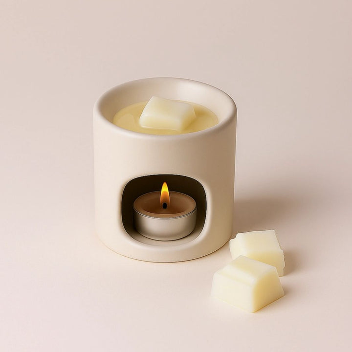 A cream-colored ceramic candle warmer holding a lit tealight candle, with pale wax cubes placed inside and beside it on a soft beige background.